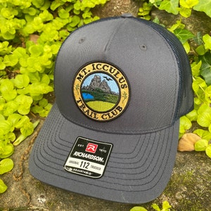 May include: A gray trucker hat with a navy blue mesh back. The hat features a circular patch with the text "MT. ICCULUS TRAIL CLUB" around an image of a mountain, lake, and green landscape. The hat has a Richardson 112 Trucker tag.
