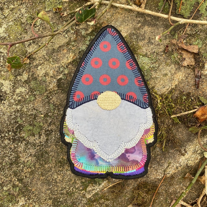 May include: A whimsical gnome patch with a blue hat featuring red circles, a gray beard, and a colorful, iridescent bottom. The patch is outlined in black and has a gold-colored nose. This decorative patch is perfect for adding a touch of fantasy to clothing or accessories.