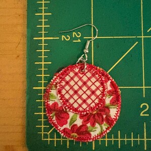May include: A handmade fabric earring with a silver hook. The earring features two circular layers of fabric. The top layer has a red and white gingham pattern, while the bottom layer displays a floral design with red flowers on a white background.