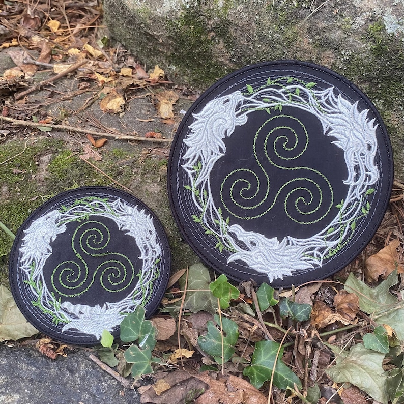 May include: Two black circular patches with embroidered designs. Each patch features a central green triple spiral, surrounded by a silver wolf design and green vines. The patches are set on a natural outdoor background.