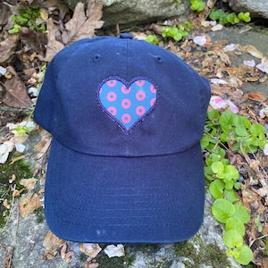May include: Navy blue baseball cap with a heart-shaped patch. The patch features a pink heart with blue donut-shaped patterns. The cap is made of a durable fabric and has a curved brim. A stylish accessory for casual wear.