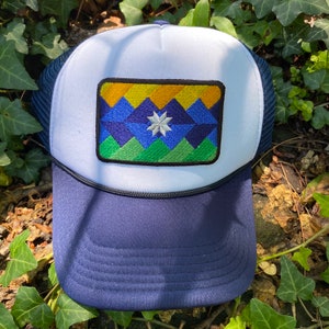 May include: A navy blue and white trucker hat with a geometric patch featuring a mountain design in shades of blue, green, and yellow. The patch includes a white star. The hat has a mesh back and a curved brim.
