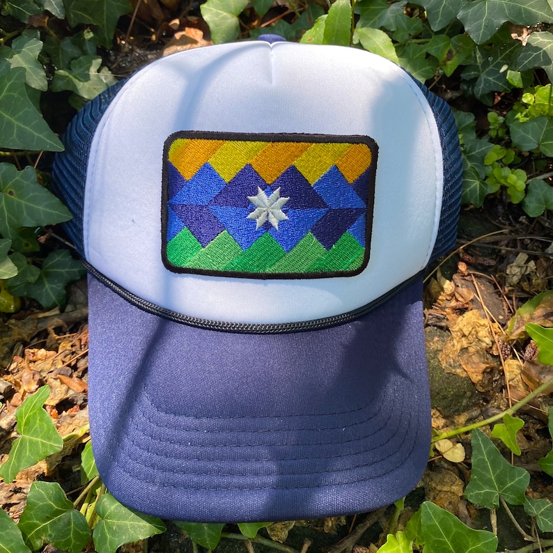 May include: A navy blue and white trucker hat with a geometric patch featuring a mountain design in shades of blue, green, and yellow. The patch includes a white star. The hat has a mesh back and a curved brim.