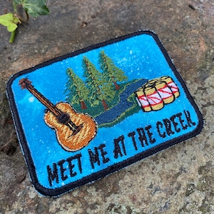 May include: Embroidered patch with a blue background featuring a guitar, trees, and a river. The patch reads "MEET ME AT THE CREEK" in blue text with a black border. The guitar is brown and the trees are green.