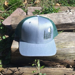 May include: A gray trucker hat with a forest scene embroidered on the front. The design features a full moon and several pine trees. The hat has a green mesh back and a curved brim.