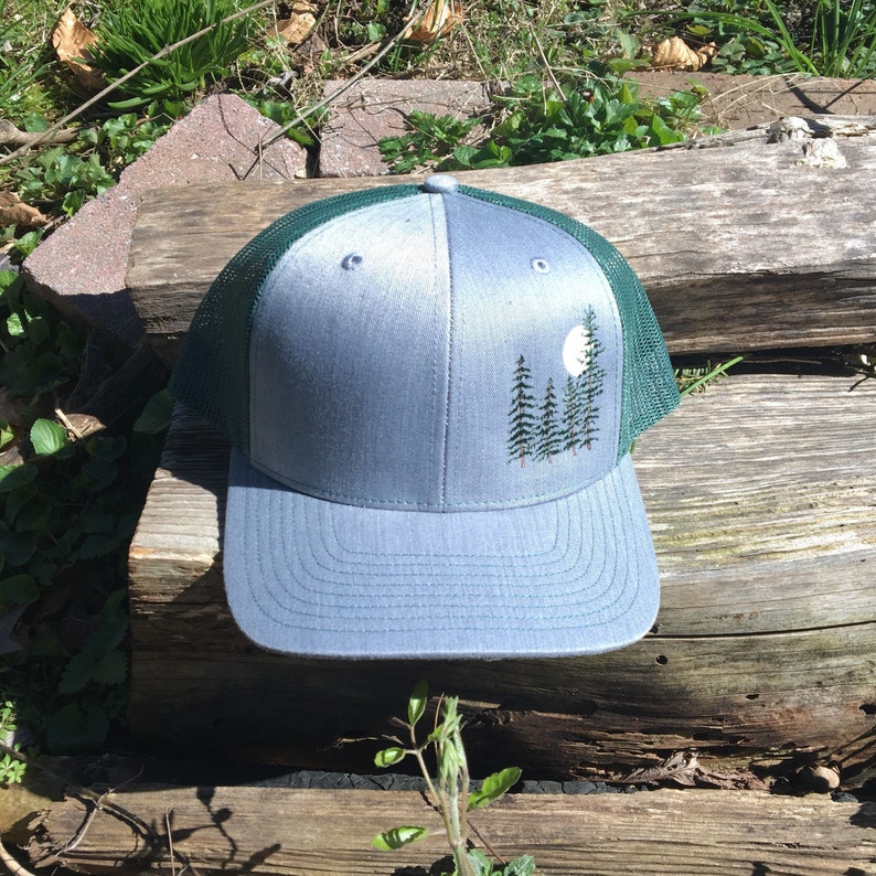 May include: A gray trucker hat with a forest scene embroidered on the front. The design features a full moon and several pine trees. The hat has a green mesh back and a curved brim.