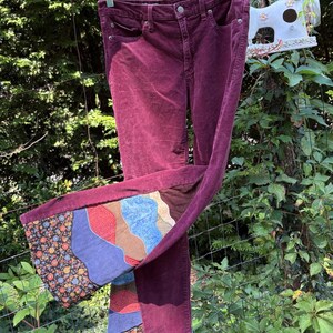 Op de afbeelding: Bordeauxkleurige corduroy broek met een patchwork ontwerp op de uitlopende zoom. Het patchwork heeft bloemen-, blauwe, rode en bruine patronen. De broek hangt buiten aan een kledinghanger.