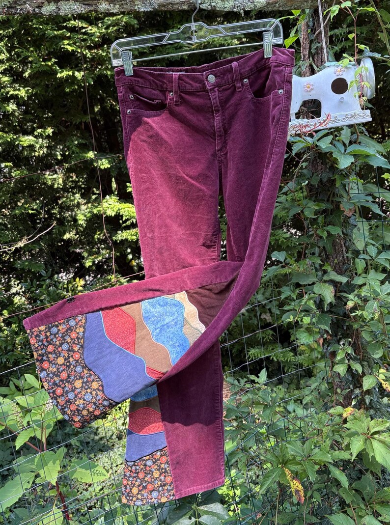 Op de afbeelding: Bordeauxkleurige corduroy broek met een patchwork ontwerp op de uitlopende zoom. Het patchwork heeft bloemen-, blauwe, rode en bruine patronen. De broek hangt buiten aan een kledinghanger.