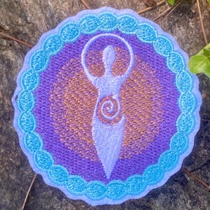 May include: Embroidered patch featuring a stylized figure with arms raised, encircled by a halo. The design is in shades of purple, gold, and light blue, with a scalloped border. The patch is circular.