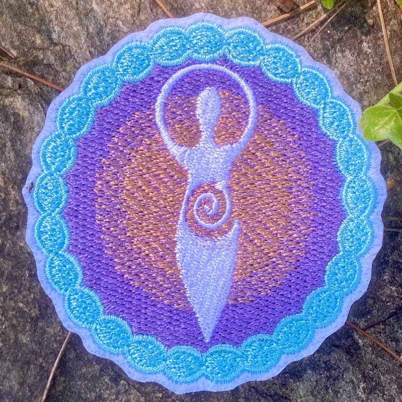May include: Circular embroidered patch with a stylized figure design. The figure is in light blue, surrounded by a purple and gold background, and a light blue scalloped border. The patch is approximately 8 cm in diameter.