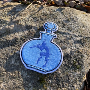 May include: A light blue embroidered patch in the shape of a potion bottle. The bottle contains a blue silhouette of a leaping figure. The patch has a white border and a gem-like design on the stopper.