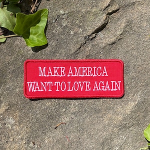 Może przedstawiać: Prostokątna czerwona naszywka z białym haftowanym napisem: "MAKE AMERICA WANT TO LOVE AGAIN." Naszywka umieszczona jest na teksturowanej szarej kamiennej powierzchni, z zielonymi liśćmi w tle.