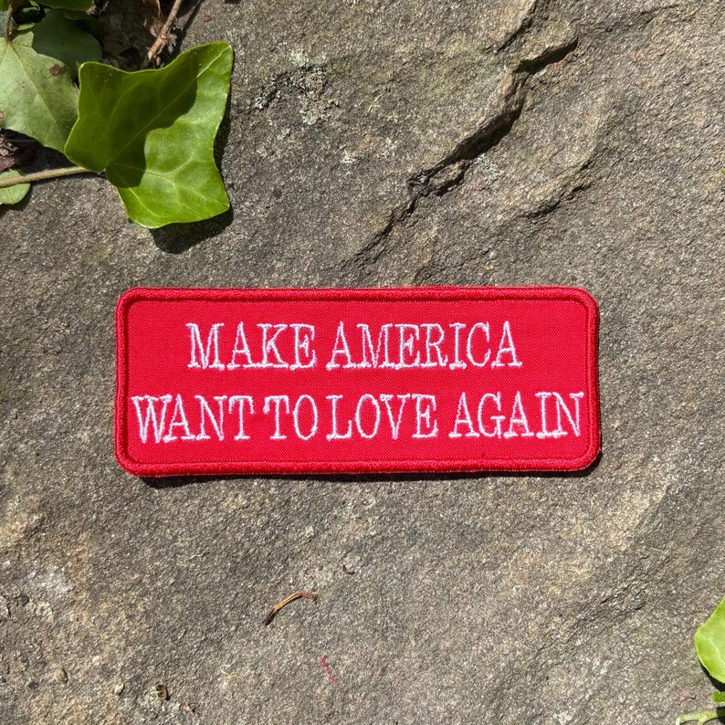 Może przedstawiać: Prostokątna czerwona naszywka z białym haftowanym napisem: "MAKE AMERICA WANT TO LOVE AGAIN." Naszywka umieszczona jest na teksturowanej szarej kamiennej powierzchni, z zielonymi liśćmi w tle.