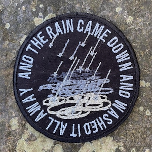 Pode incluir: Remendo redondo bordado preto com texto branco: "AND THE RAIN CAME DOWN AND WASHED IT ALL AWAY". O design inclui chuva e nuvens bordadas em tons de cinza e branco.