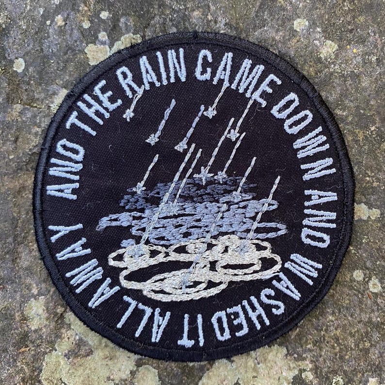 Pode incluir: Remendo redondo bordado preto com texto branco: "AND THE RAIN CAME DOWN AND WASHED IT ALL AWAY". O design inclui chuva e nuvens bordadas em tons de cinza e branco.