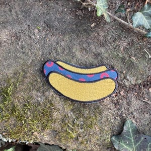 May include: Embroidered patch in the shape of a hot dog. The hot dog bun is yellow with a black outline. The hot dog has a blue stripe with red polka dots. The patch is on a stone surface.