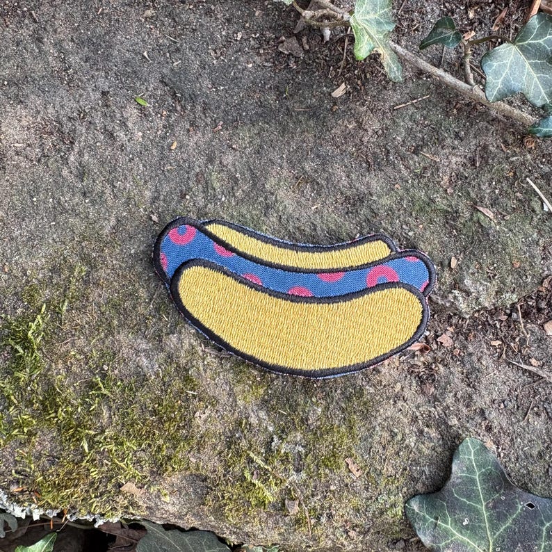 May include: Embroidered patch in the shape of a hot dog. The hot dog bun is yellow with a black outline. The hot dog has a blue stripe with red polka dots. The patch is on a stone surface.