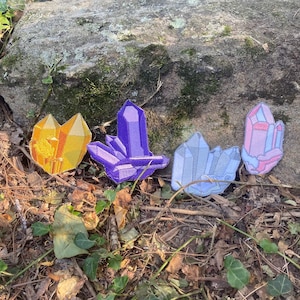 May include: Four embroidered crystal patches in shades of gold, purple, light blue, and pink. The patches are arranged on a bed of leaves and dirt, with a large rock in the background. The crystals have geometric shapes and are likely for decorative purposes.