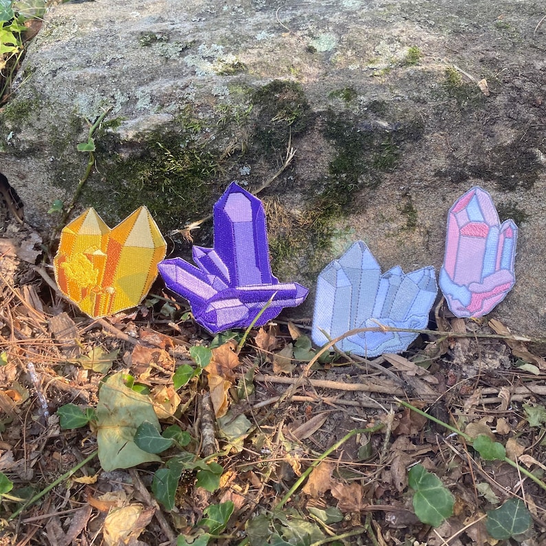 May include: Four embroidered crystal patches in gold, purple, light blue, and pink. The patches are arranged on leaves and dirt, with a large rock behind them. The crystals have geometric shapes and are likely for decorative purposes.