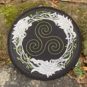 May include: Round black patch with white embroidered wolf heads and green vines encircling a green spiral design. The patch has a stitched border and is set against a natural, mossy background.
