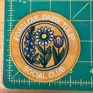 Pu&ograve; includere: Toppa rotonda ricamata color giallo senape con la scritta bianca "FOXGLOVE DAISY TULIP SOCIAL CLUB". Presenta illustrazioni di una margherita, un tulipano e una digitale, nei toni del viola, rosa e bianco.