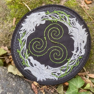 May include: Round black embroidered patch featuring a stylized wolf design in gray and green. The patch has a central triskele symbol in green, surrounded by a wreath of green vines and two wolf heads.