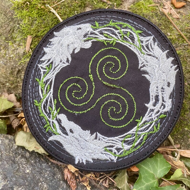 May include: Round black embroidered patch featuring a stylized wolf design in gray and green. The patch has a central triskele symbol in green, surrounded by a wreath of green vines and two wolf heads.