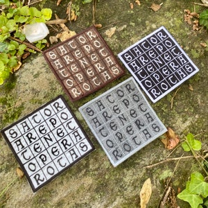 May include: Four square embroidered patches with the text "SATOR AREPO TENET OPERA ROTAS" in a grid pattern. The patches are in brown, white, black, and gray, with the text in contrasting colors. The patches are displayed on a natural, green surface.