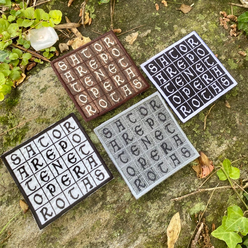 May include: Four square embroidered patches with the text "SATOR AREPO TENET OPERA ROTAS" in a grid pattern. The patches are in brown, white, black, and gray, with the text in contrasting colors. The patches are displayed on a natural, green surface.