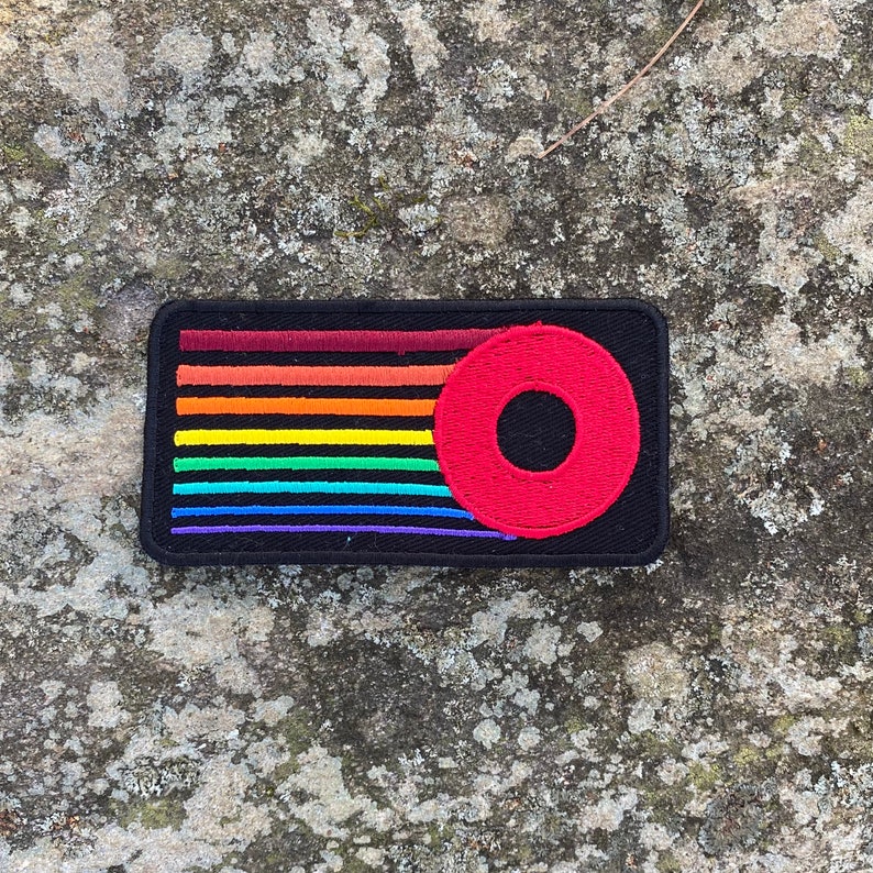 K&ouml;nnte beinhalten: Ein rechteckiger schwarzer Aufn&auml;her mit einem Regenbogenstreifen-Design und einem roten Kreis mit einem Loch. Die Regenbogenstreifen sind horizontal und umfassen Rot, Orange, Gelb, Gr&uuml;n, Blau und Lila. Es ist wahrscheinlich ein gesticktes Design.