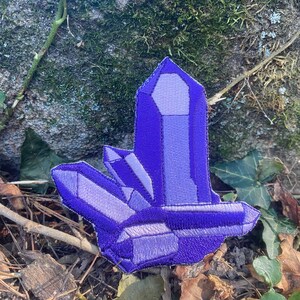 May include: A purple crystal patch with embroidered details. The patch features a cluster of crystals in varying shades of purple, with a detailed, textured appearance. The patch is set against a natural outdoor background.