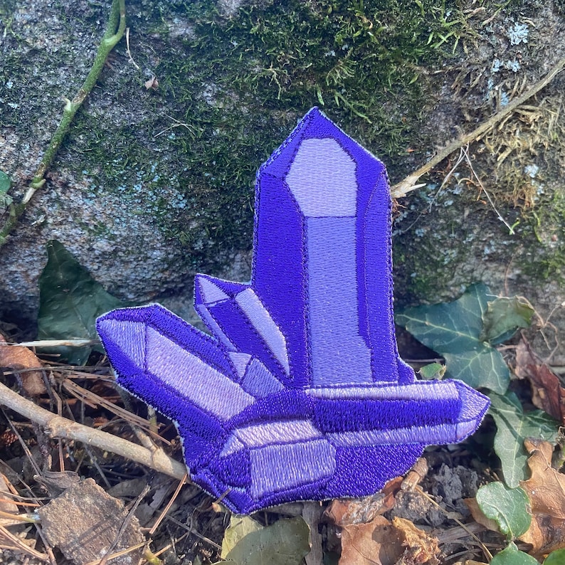 May include: A purple crystal patch with embroidered details. The patch features a cluster of crystals in varying shades of purple, with a detailed, textured appearance. The patch is set against a natural outdoor background.