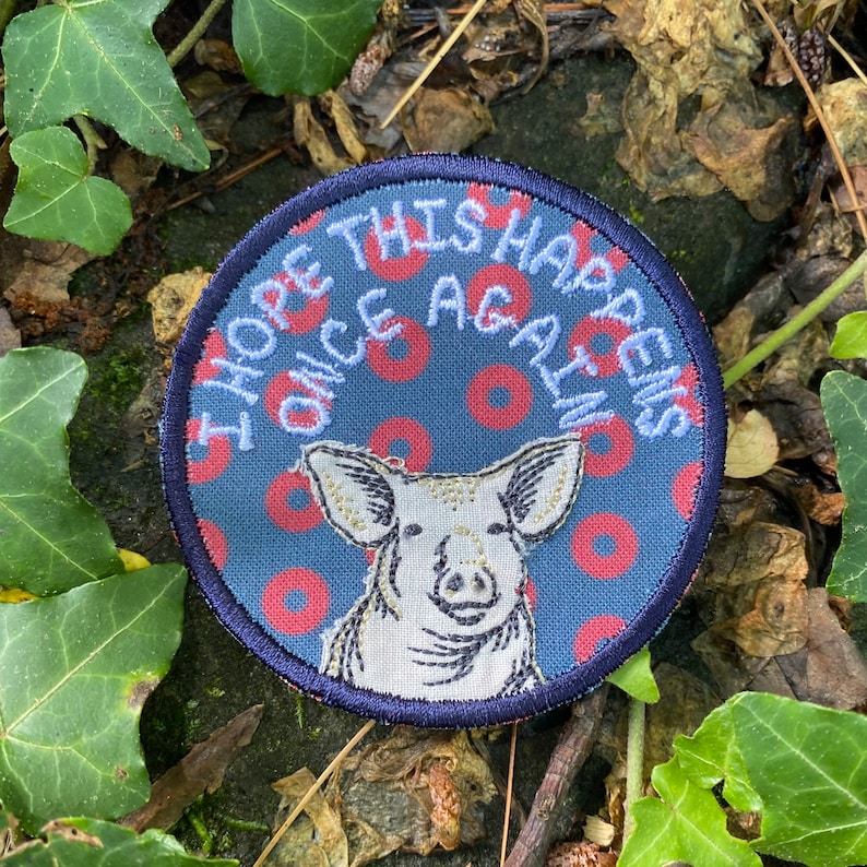 May include: Round patch with a navy blue border and background featuring a pig illustration. The patch has the text "I HOPE THIS HAPPENS ONCE AGAIN" in white. The background has a pattern of red circles.