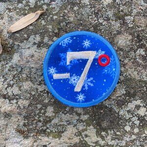 Op de afbeelding: Een ronde, blauwe patch met witte sneeuwvlokken en het getal -7 in het wit. Een kleine rode cirkel staat naast het getal 7. De patch ligt op een getextureerd grijs stenen oppervlak.