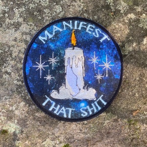 May include: Round patch with a blue galaxy background and the words "MANIFEST THAT SHIT" in white. A white candle with a yellow flame is in the center, surrounded by white starbursts. The patch has a black border.