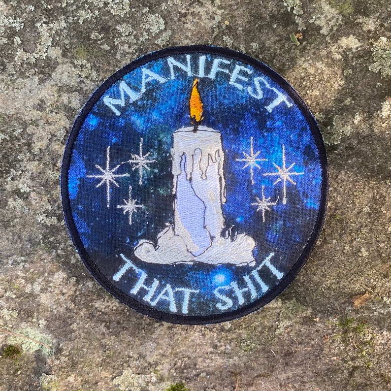 May include: Round patch with a blue galaxy background and the words "MANIFEST THAT SHIT" in white. A white candle with a yellow flame is in the center, surrounded by white starbursts. The patch has a black border.