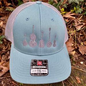 Pode incluir: Bon&eacute; trucker Richardson 115 turquesa com parte traseira em malha cinza. A frente do bon&eacute; apresenta bordados cinza de instrumentos de cordas. O bon&eacute; tem uma aba curva e fecho de encaixe. O logotipo da Richardson &eacute; vis&iacute;vel.