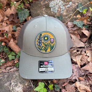 May include: A green trucker hat with a floral patch featuring a daisy, tulip, and other flowers. The hat has a mesh back and a curved brim. The Richardson brand label is visible on the front.
