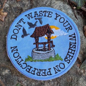May include: Round blue embroidered patch with the text "DON'T WASTE YOUR WISHES ON PERFECTION". The patch features a well with a brown roof, two birds, a yellow sun, and white clouds.