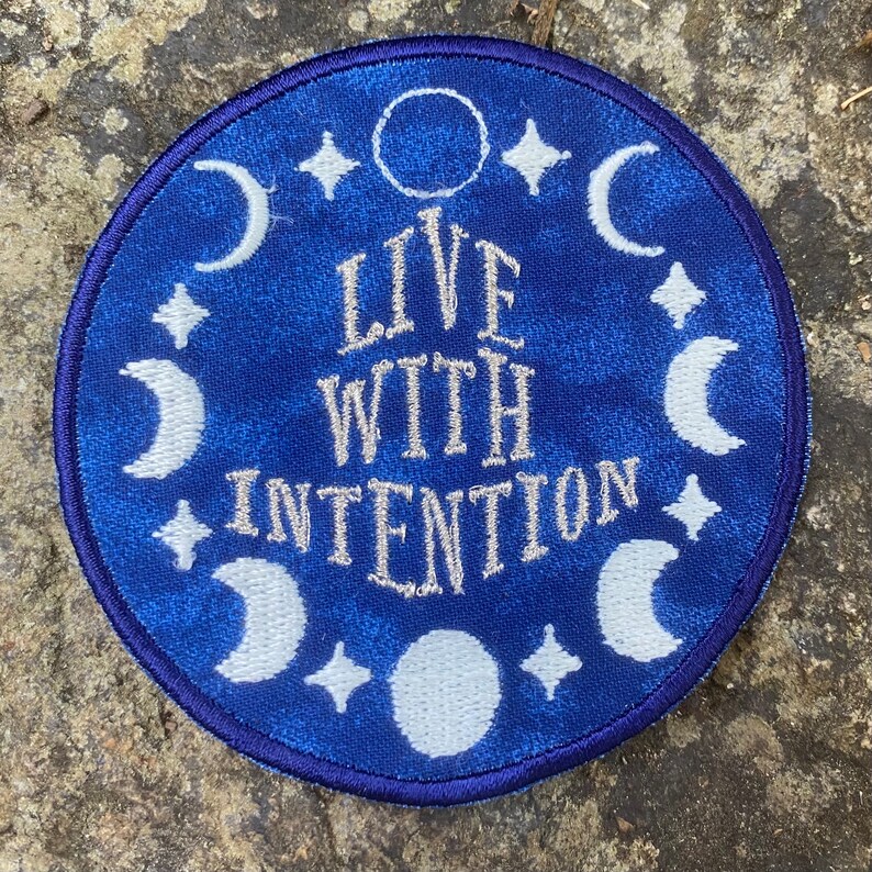 May include: A circular, blue embroidered patch with the words "LIVE WITH INTENTION" in white script. The patch features white crescent moons, stars, and a full moon design around the edge. The patch has a dark blue border.