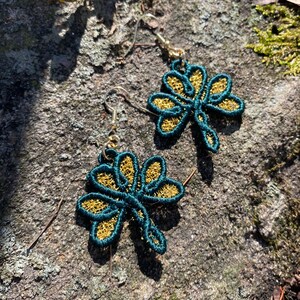 Pode incluir: Um par de brincos de lib&eacute;lula em verde-azulado e dourado. Os brincos apresentam um contorno verde-azulado com asas de filigrana dourada. Os brincos penduram de ganchos dourados.