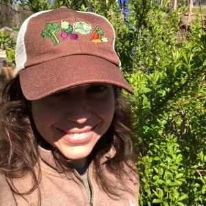 May include: A brown baseball cap with a white mesh back. The front of the cap features embroidered vegetables: broccoli, beets, cabbage, and a carrot. The cap is worn outdoors.