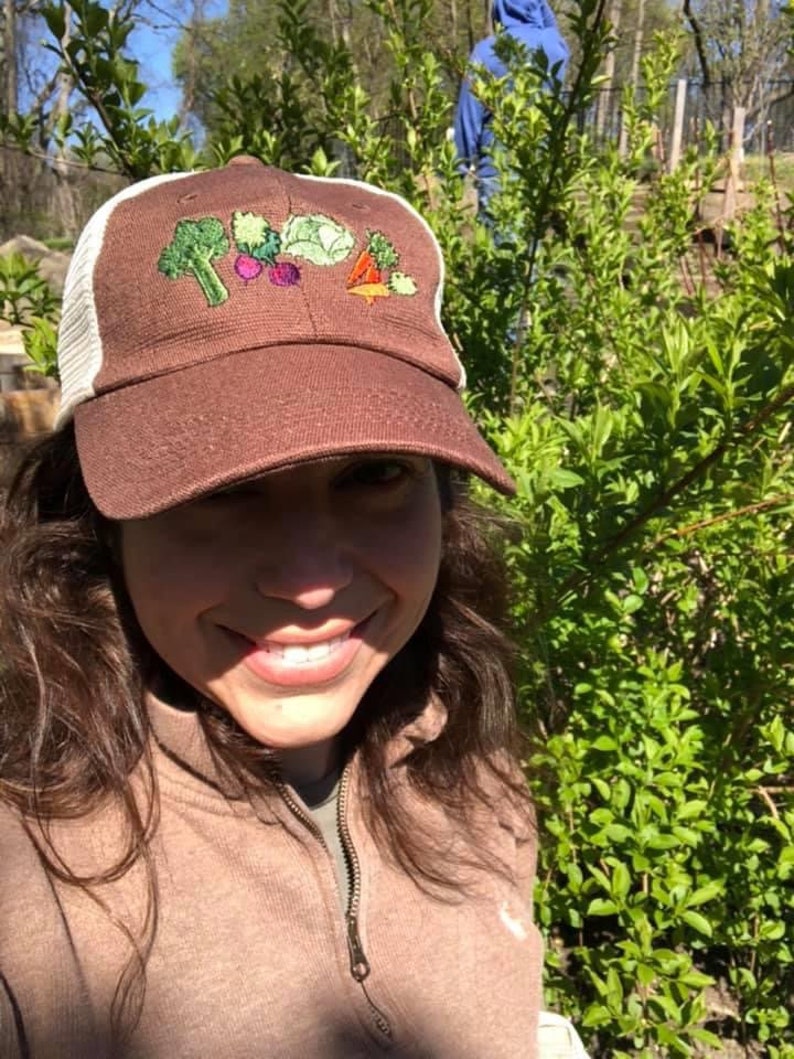 May include: A brown baseball cap with a white mesh back. The front of the cap features embroidered vegetables: broccoli, beets, cabbage, and a carrot. The cap is worn outdoors.