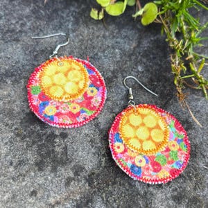 Pu&ograve; includere: Un paio di orecchini in tessuto fatti a mano. Ogni orecchino presenta un design circolare con un centro giallo e un vivace motivo floreale colorato su sfondo rosso. Gli orecchini hanno ganci argentati.