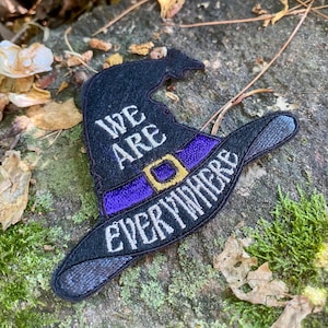 May include: Black witch hat patch with the words "WE ARE EVERYWHERE" embroidered in white. The hat features a purple band with a gold buckle. The patch is placed on a mossy rock.