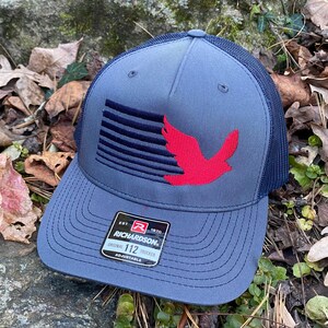 May include: A gray trucker hat with a navy blue mesh back and brim. The front panel features a red bird design and a navy blue striped design. The hat has the text "RICHARDSON" and "ORIGINAL 112 TRUCKER" on a tag.