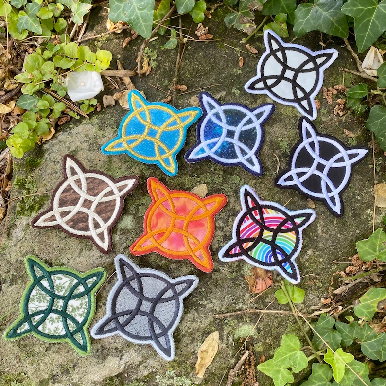 May include: A collection of embroidered patches featuring a Celtic knot design. The patches display various colors and patterns, including blue, black, brown, and rainbow. The patches are arranged on a stone surface with greenery.