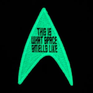 May include: A neon green embroidered patch in the shape of a triangle with rounded edges. The patch has the text "THIS IS WHAT SPACE SMELLS LIKE" in black, block letters. The background is solid black.