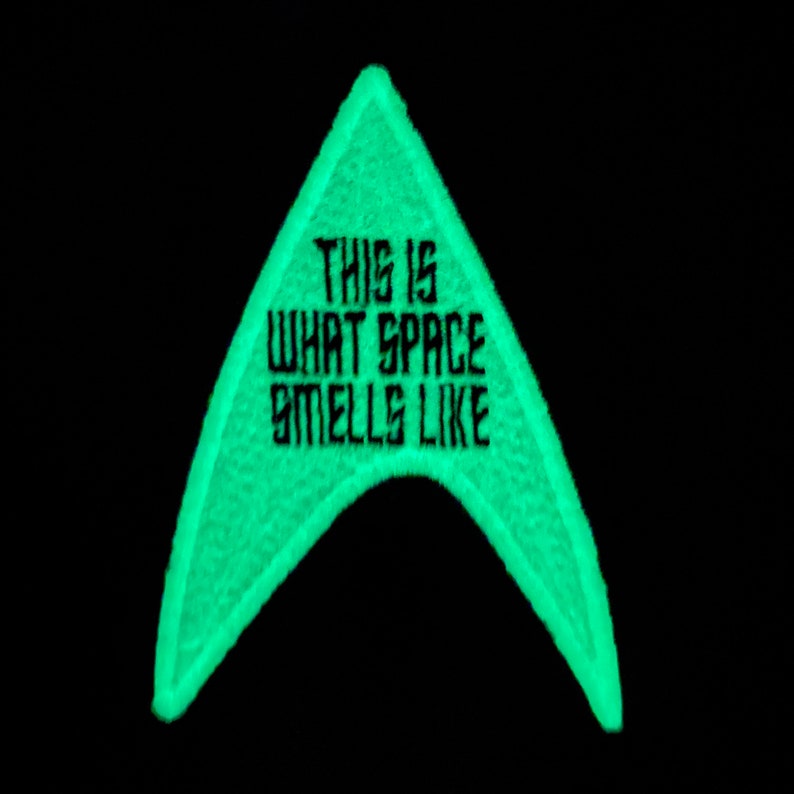 May include: A neon green embroidered patch in the shape of a triangle with rounded edges. The patch has the text "THIS IS WHAT SPACE SMELLS LIKE" in black, block letters. The background is solid black.