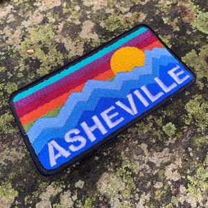 May include: Embroidered patch featuring a colorful mountain and sunset design with the word "ASHEVILLE" in white. The patch has a black border and is rectangular. The background is a mossy rock.